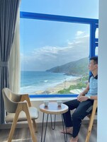 Una habitación con un gran ventanal que da a una playa, una silla, una pequeña mesa con dos tazas, y una persona sentada junto a la ventana.