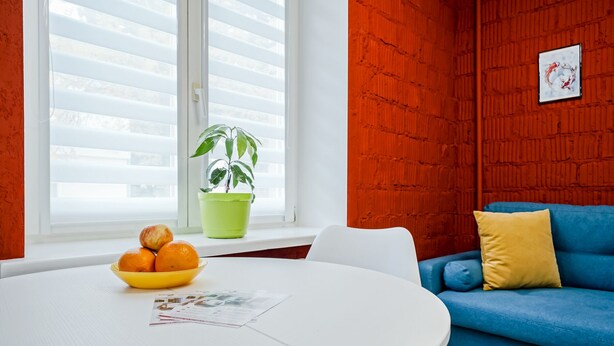 Una habitación con una mesa redonda blanca, un sofá azul con cojines amarillos y azules, una planta en maceta verde y una ventana con persianas.