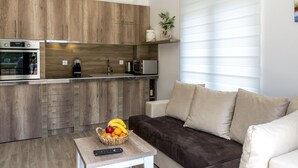 A modern kitchen with wooden cabinets, a built-in oven, and a coffee machine. A basket of fruit is on a small table, and a sofa is in the living area with a window. - Stylish Apartment With Balcony & Pool Access Near The Historical Town (Kotor)