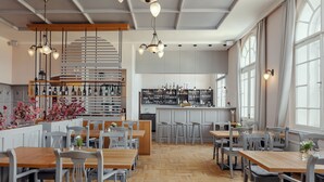 Un restaurante con mesas y sillas de madera, una zona de bar con estantes llenos de botellas y ventanas amplias que dejan entrar luz natural.
