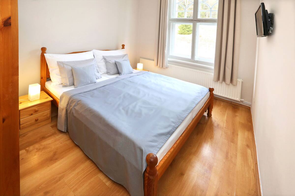 A neatly made bed with a wooden frame and white bedding, a bedside table with a lamp, and a window with curtains.