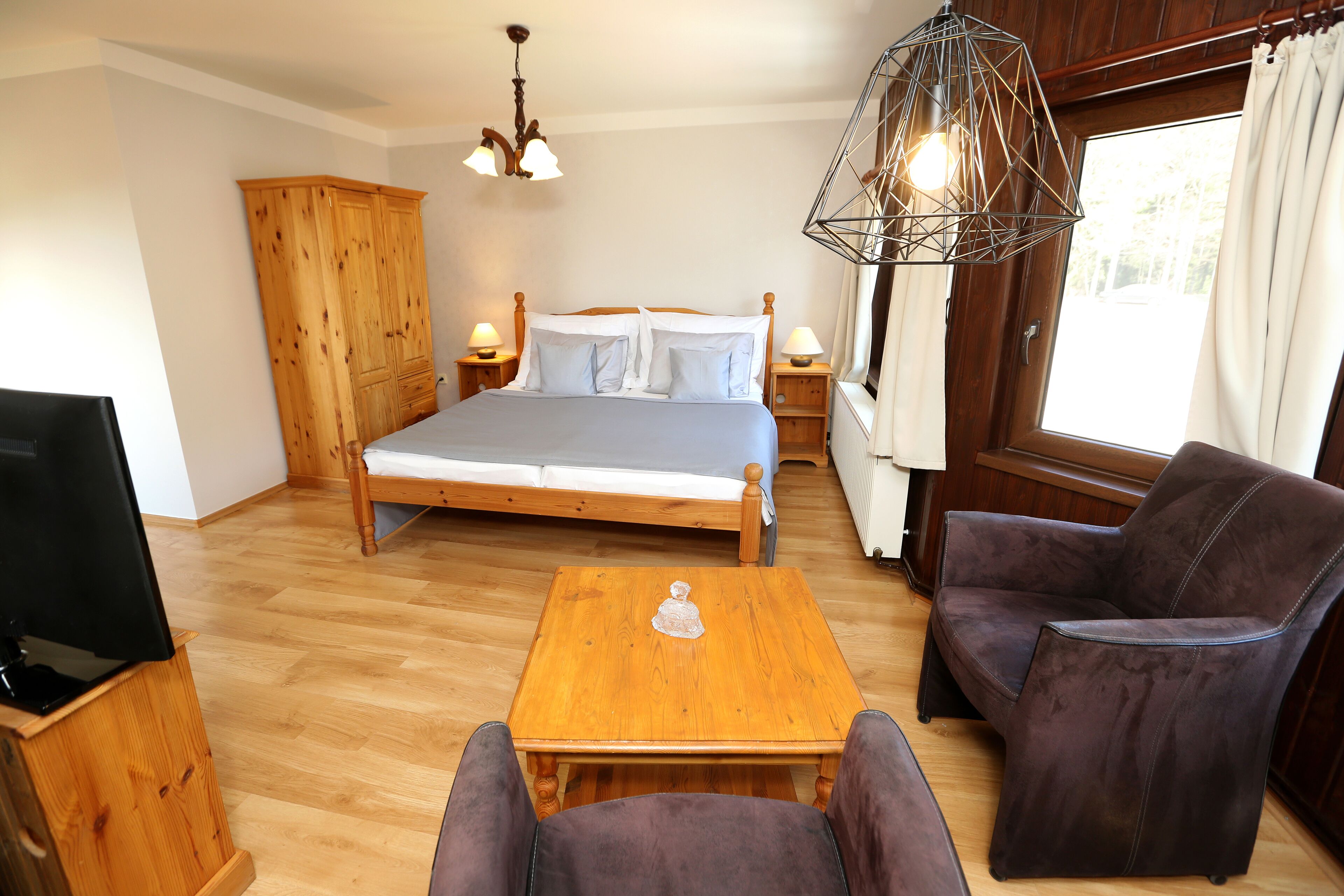 Quarto com cama, guarda-roupa de madeira, duas mesas de cabeceira com abajures, uma mesa de centro de madeira e duas poltronas.