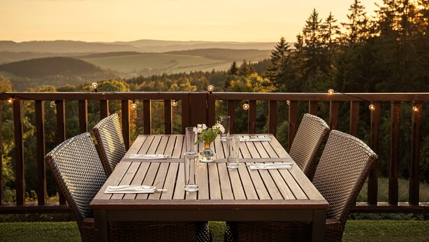 Una terraza de madera con una mesa dispuesta para comer, con vistas a un paisaje pintoresco de colinas y árboles.