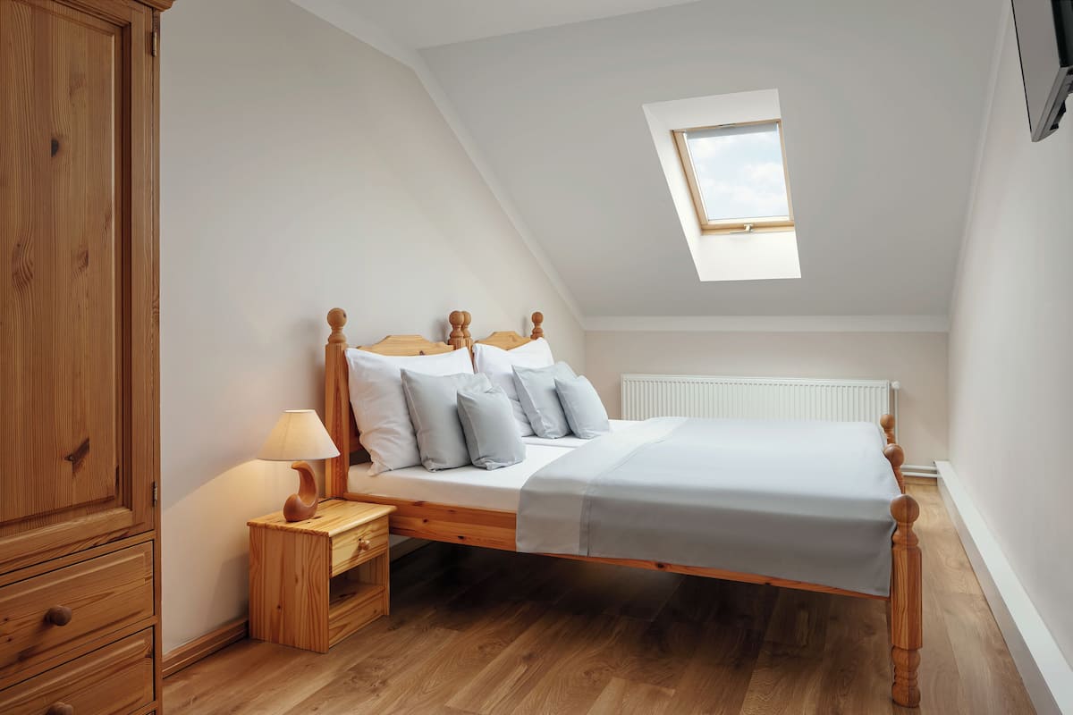 A bedroom with a sloped ceiling, a skylight, a wooden bed frame, a bedside table with a lamp, and a wooden wardrobe.