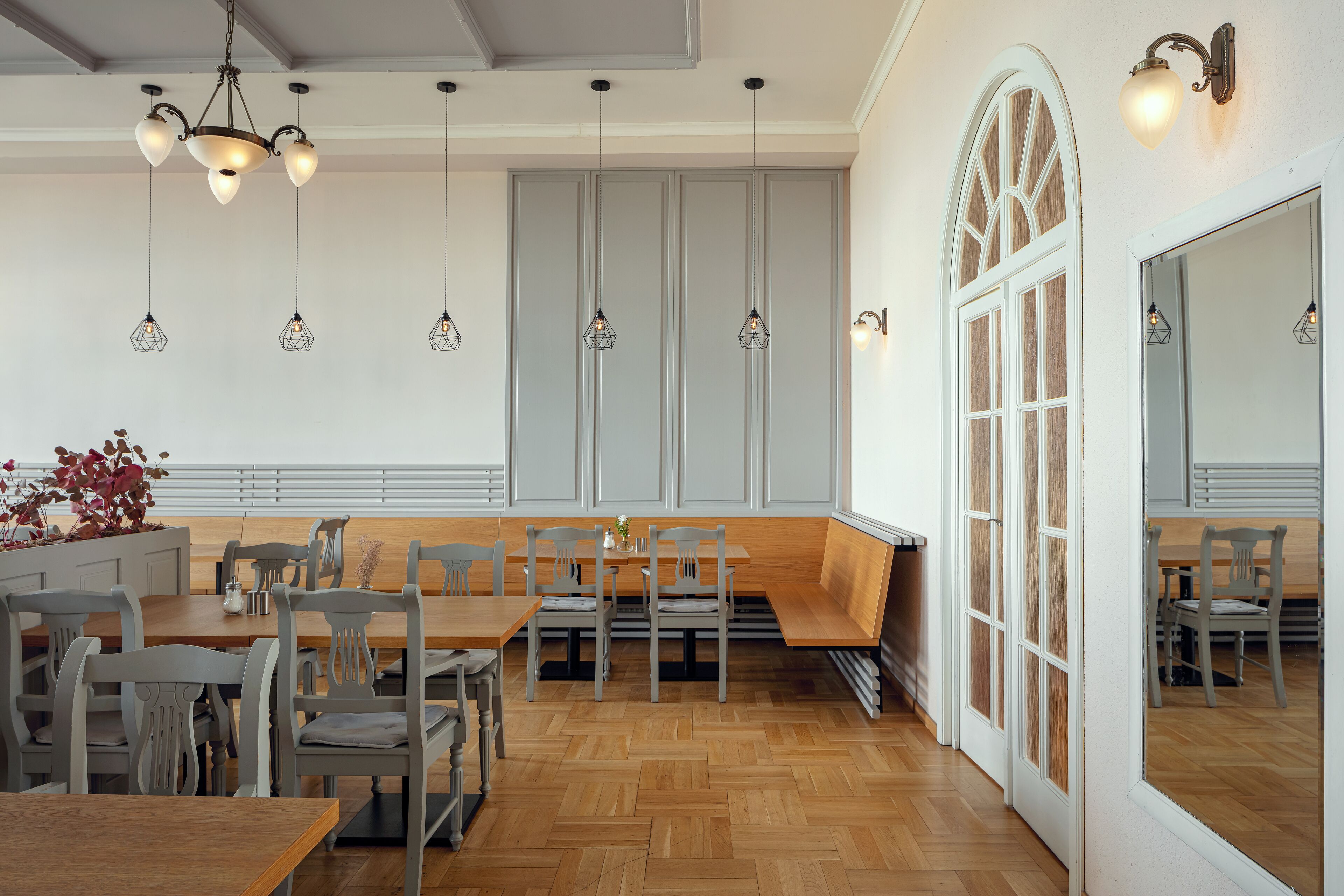 A dining area with wooden tables and chairs, hanging pendant lights, and a large arched window.