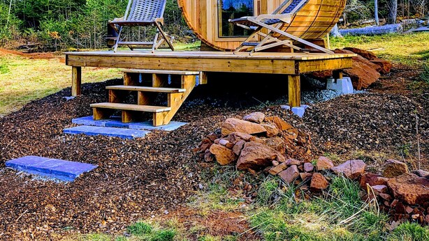 A wooden cabin with a large barrel-shaped door, two lounge chairs, and a small table with a solar panel.