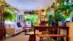 A cozy living room with a sofa, wooden coffee table, and a bench. The room is decorated with numerous potted plants and framed artwork.