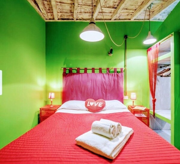 Una habitación con paredes verdes, una colcha roja, un cojín con forma de corazón que dice 'LOVE' y una toalla enrollada sobre la cama.