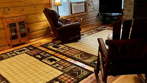 A cozy log cabin interior with a fireplace, a television, a leather armchair, and a patterned rug.