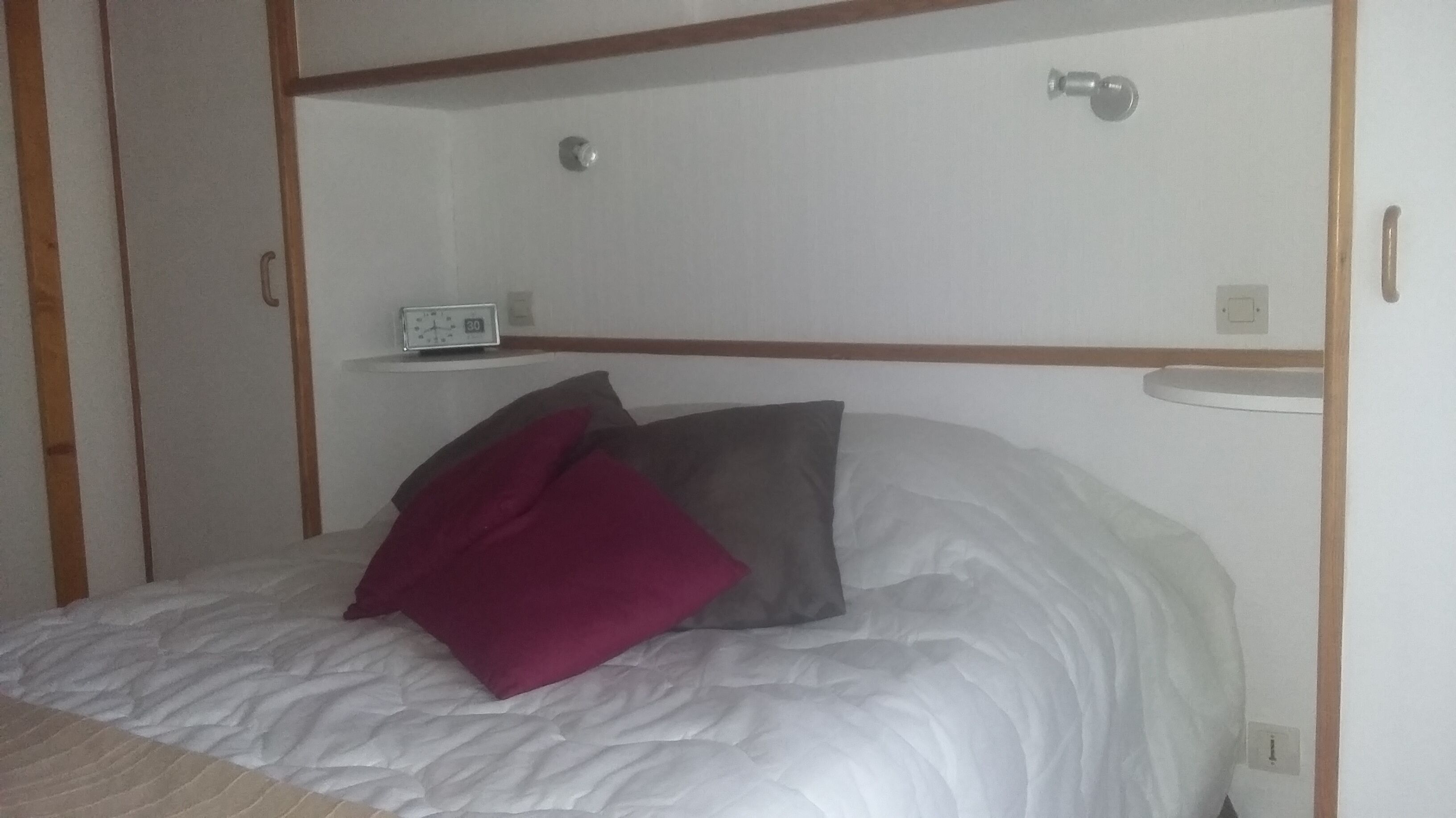 Ein Einzelbett mit weißer Matratze und zwei Kissen, eines rot und eines grau, in einem Zimmer mit Holzvertäfelung und einem kleinen Nachttisch mit einer Uhr.