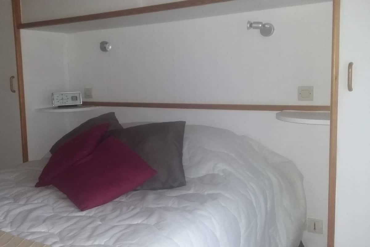 Ein Einzelbett mit weißer Matratze und zwei Kissen, eines rot und eines grau, in einem Zimmer mit Holzvertäfelung und einem kleinen Nachttisch mit einer Uhr.
