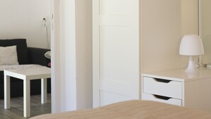 A modern bedroom with a white bed, a bedside table, a white dresser with a mirror, and a black couch with patterned cushions. - Le Hameau de la Courbière (Tarascon-sur-Ariege)