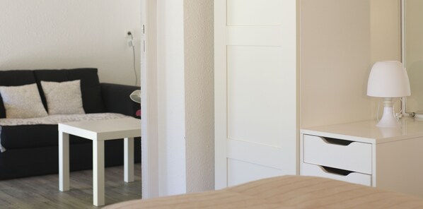 Un dormitorio moderno con una cama blanca, una mesita de noche, un tocador blanco con espejo y un sofá negro con cojines estampados.
