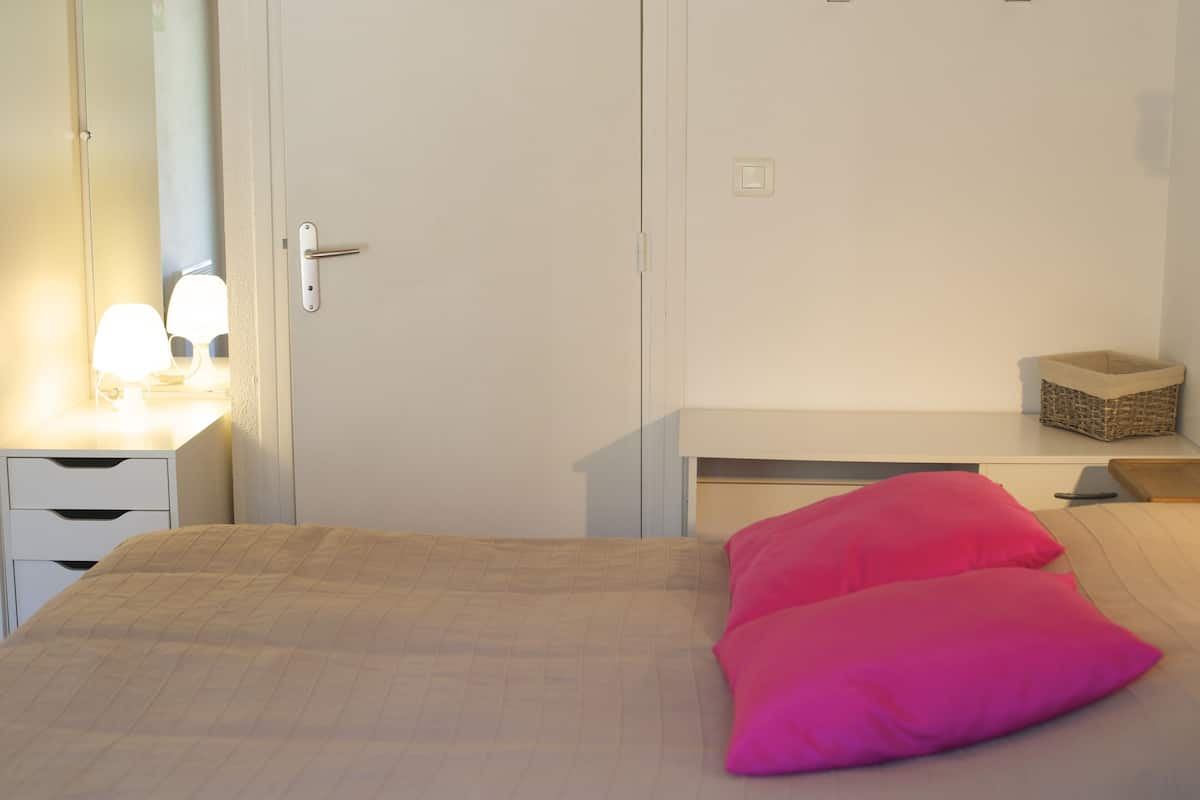 Ein Schlafzimmer mit einem Bett, einem Nachttisch mit Lampe, einer Kommode und einem kleinen Korb auf dem Nachttisch.