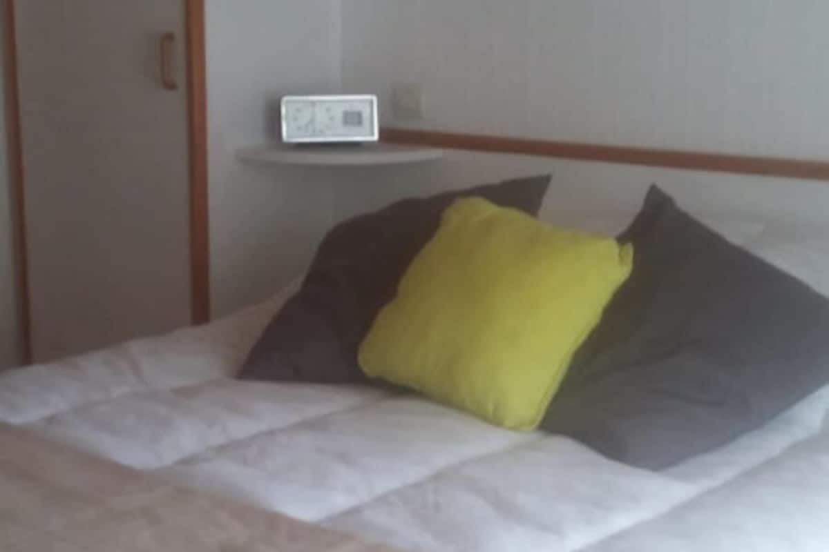 Ein Bett mit einer weißen Matratze, einem gelben Kissen und einem grauen Kissen. Darüber befindet sich eine kleine Ablagefläche mit einer Uhr.