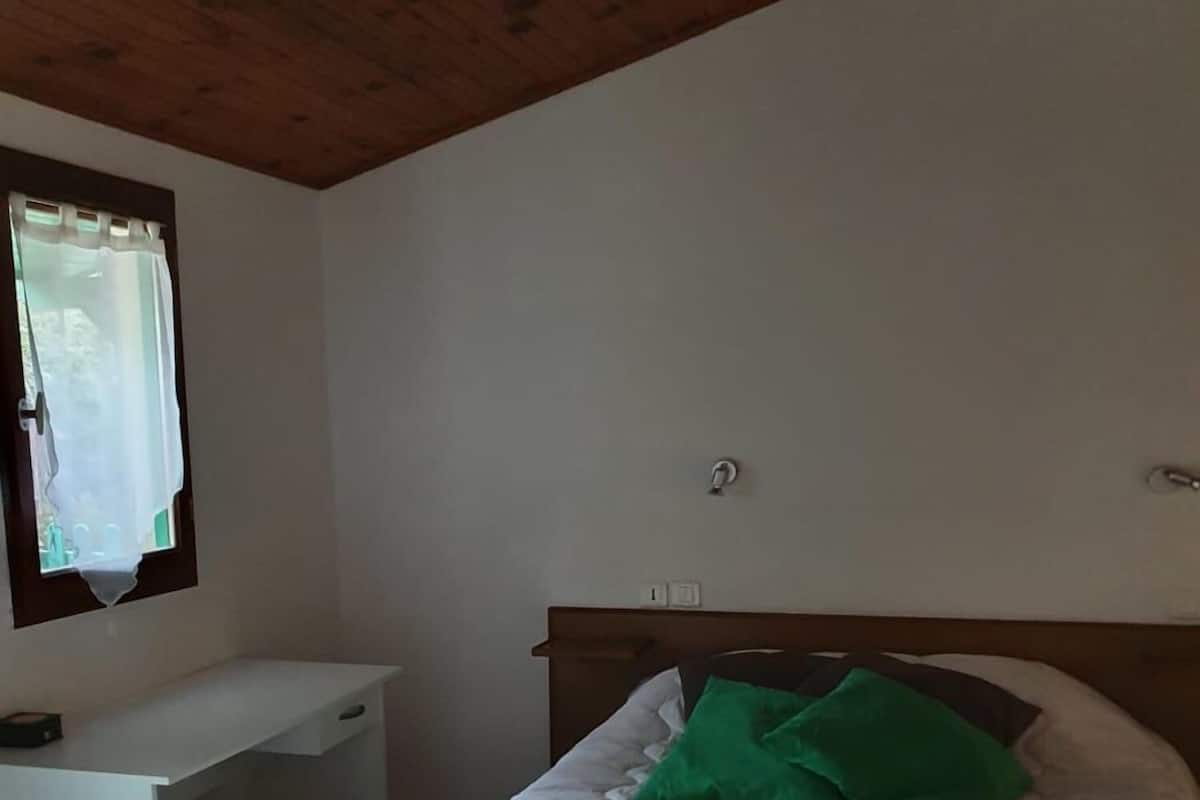 Ein Schlafzimmer mit Holzboden, einem Bett mit weißer Bettwäsche und grünen Kissen, einem weißen Schreibtisch und einem Fenster mit Vorhang.