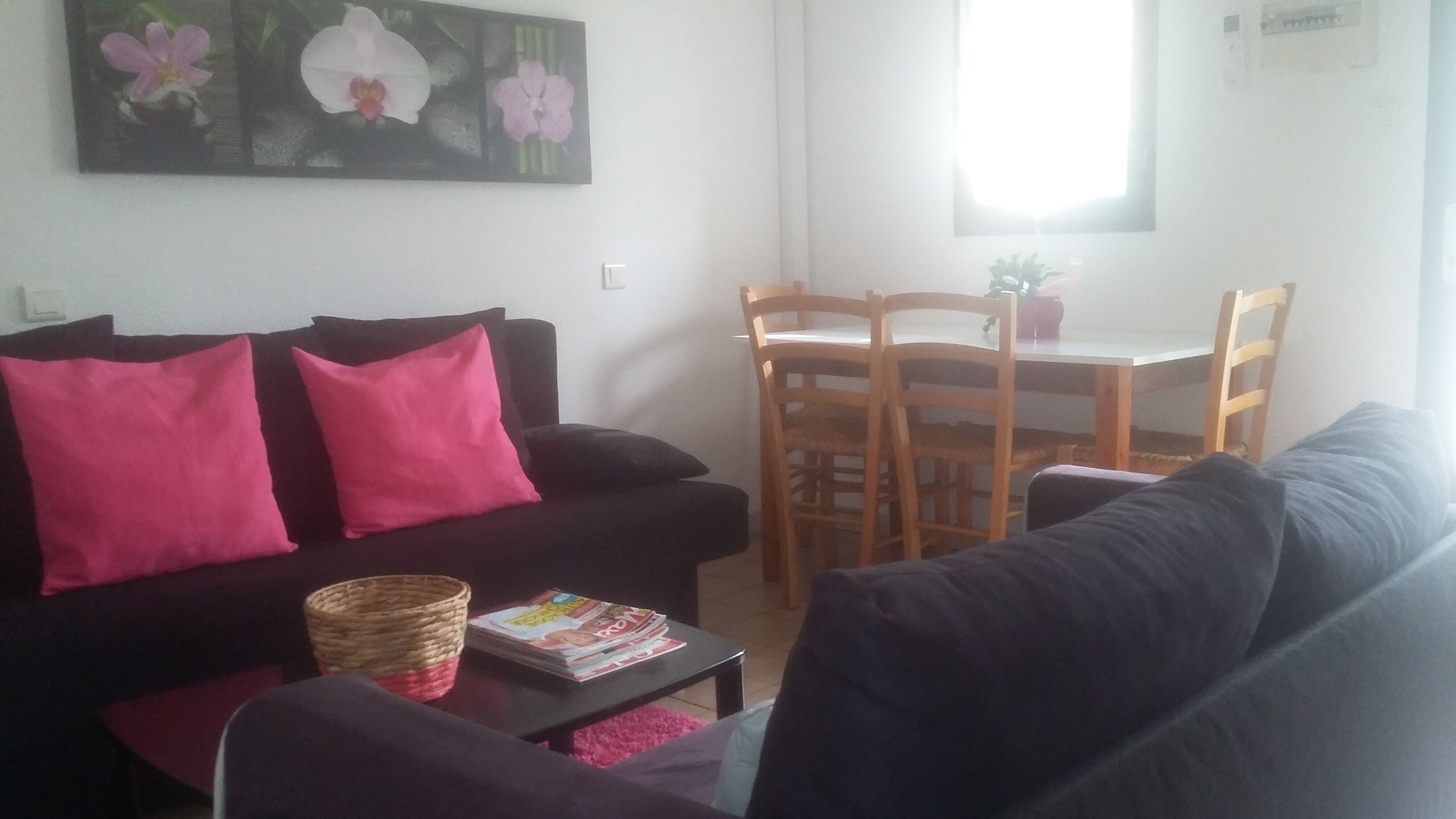 A living room with a black sofa, two pink cushions, a wicker basket, and a magazine on a small table. There is a dining table with chairs and a window in the background.