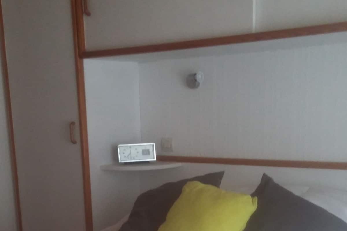 Ein Bett mit einer weißen Matratze, einem grauen Kissen und einem gelben Kissen. Ein weißer Nachttisch mit einer Uhr steht daneben.