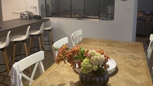 A dining area with a wooden table, white chairs, and a vase of flowers. There is a window with a view of the outside. - COMFORTABLE HOUSE CLOSE TO ALL AMENITIES (Blainville-sur-Mer)