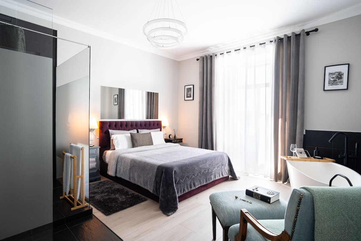 A modern hotel room with a large bed, a sitting area with a chair, a desk, a bathtub, and a large window with curtains.
