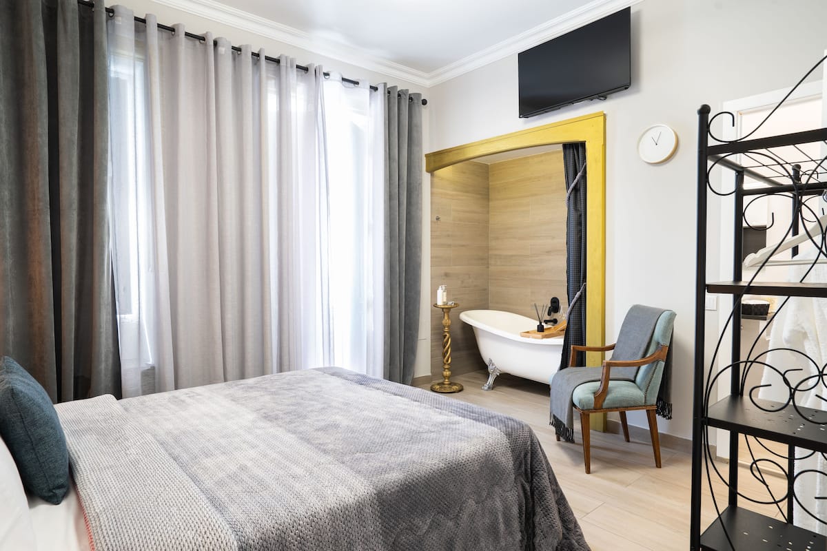 A hotel room with a bed, a chair, a bathtub, and a TV on the wall.