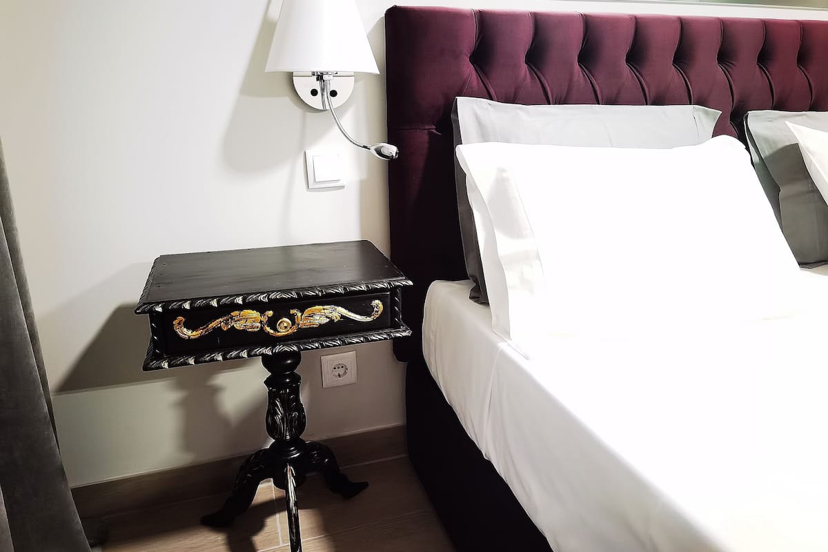 A neatly made bed with white linens, a dark wooden nightstand with a decorative top, and a wall-mounted lamp.