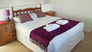 Una cama bien hecha con una manta morada, almohadas blancas y un cabecero de madera. Hay una mesita de noche de madera con una lámpara y un cajón pequeño. Un cuadro grande con flores está en la pared sobre la cama.