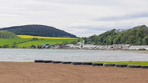 Vista a la orilla del río con un sendero de tierra, campos verdes y una hilera de casas junto al agua.