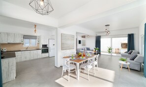 A modern kitchen with white cabinetry, a dining area with a wooden table and white chairs, and a living area with a grey sofa and a blue curtain.