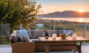 A wicker sofa with cushions, a table with a bottle of wine, glasses, and a bowl of fruit, overlooking a scenic view of a body of water and distant buildings. - Cretan Sunset Villa | Heated Pool (Georgioupoli)