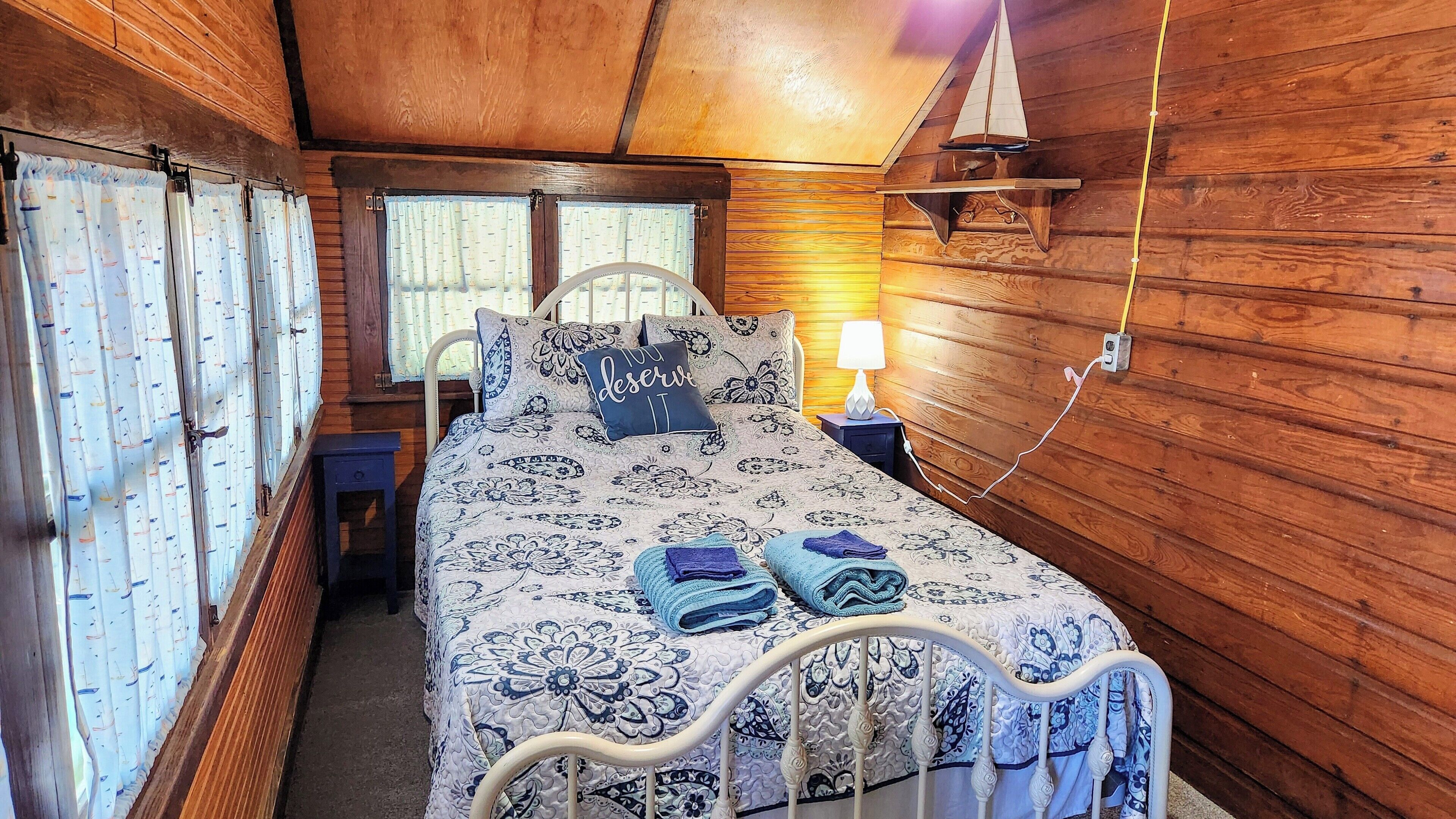 Ein Schlafzimmer einer Holzhütte mit einem Bett, Kissen, einem Nachttisch, einer Lampe und einer kleinen Segelbohrminieatur.