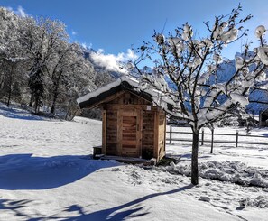 A wooden cabin with a snow-covered roof, surrounded by snow-covered trees and mountains in the background.