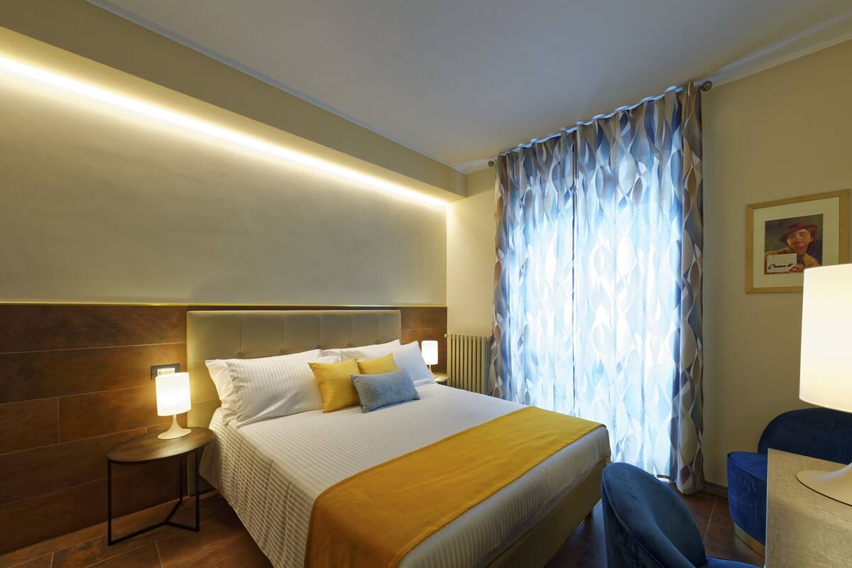 A hotel room with a large bed, a bedside table with a lamp, a chair, and a window with patterned curtains.
