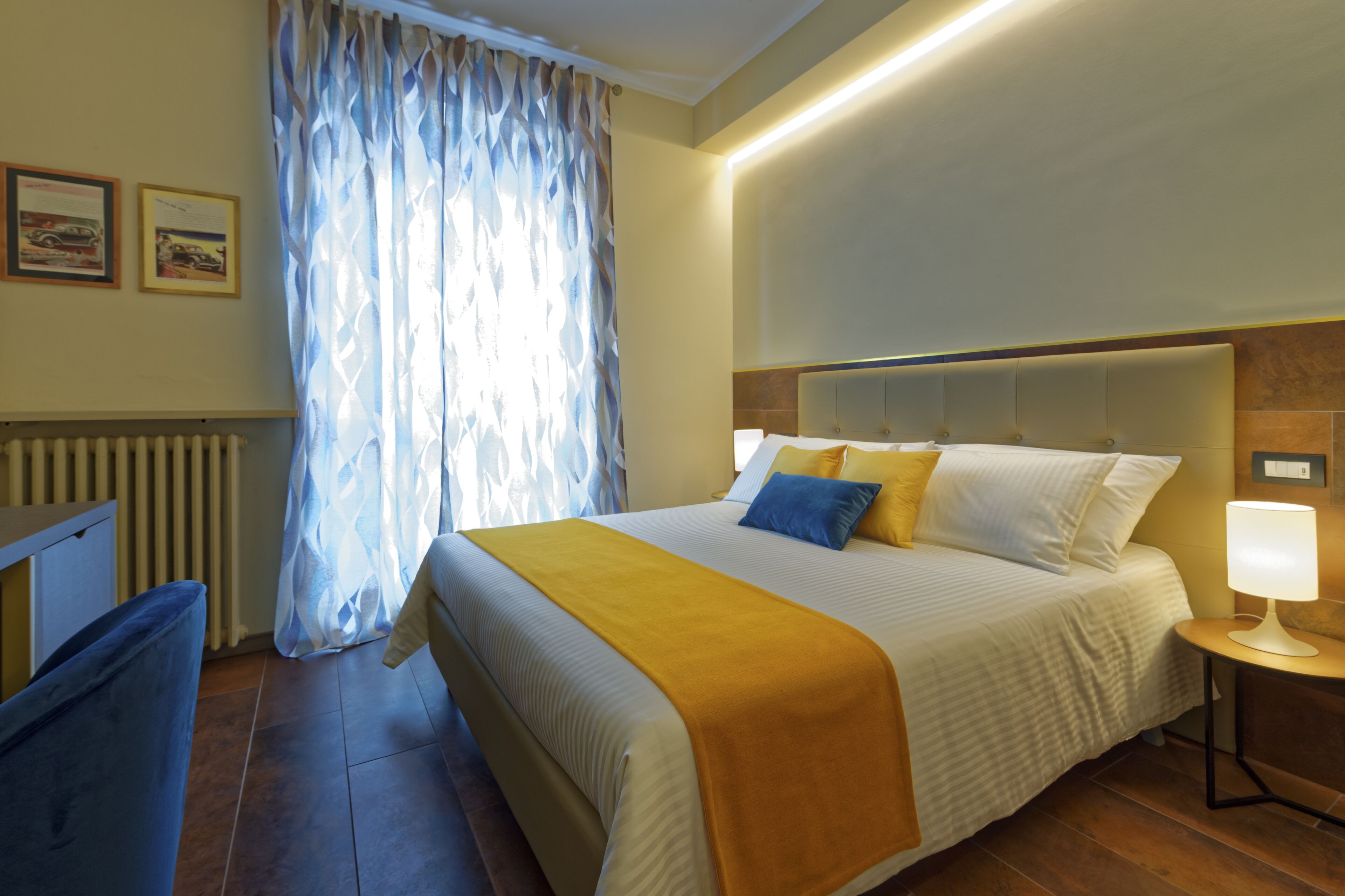 Une chambre d’hôtel avec un grand lit, une table de chevet avec une lampe, un radiateur et une fenêtre avec des rideaux.