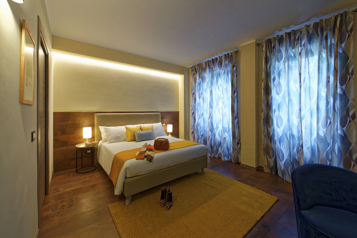 A hotel room with a large bed, a bedside table with a lamp, a chair, and a view of the outside through patterned curtains.
