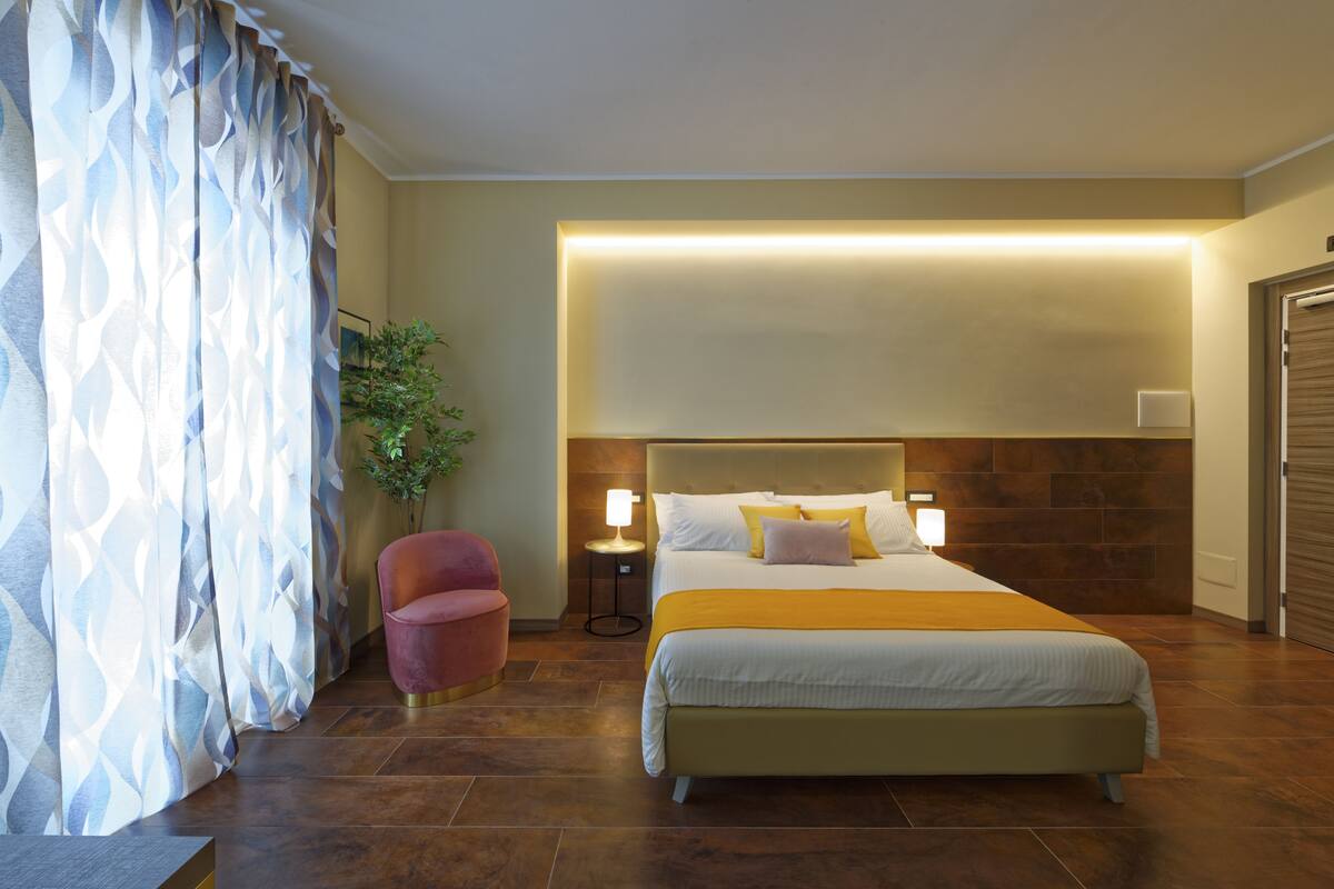 A hotel room with a large bed, a nightstand with a lamp, a potted plant, and a red chair.