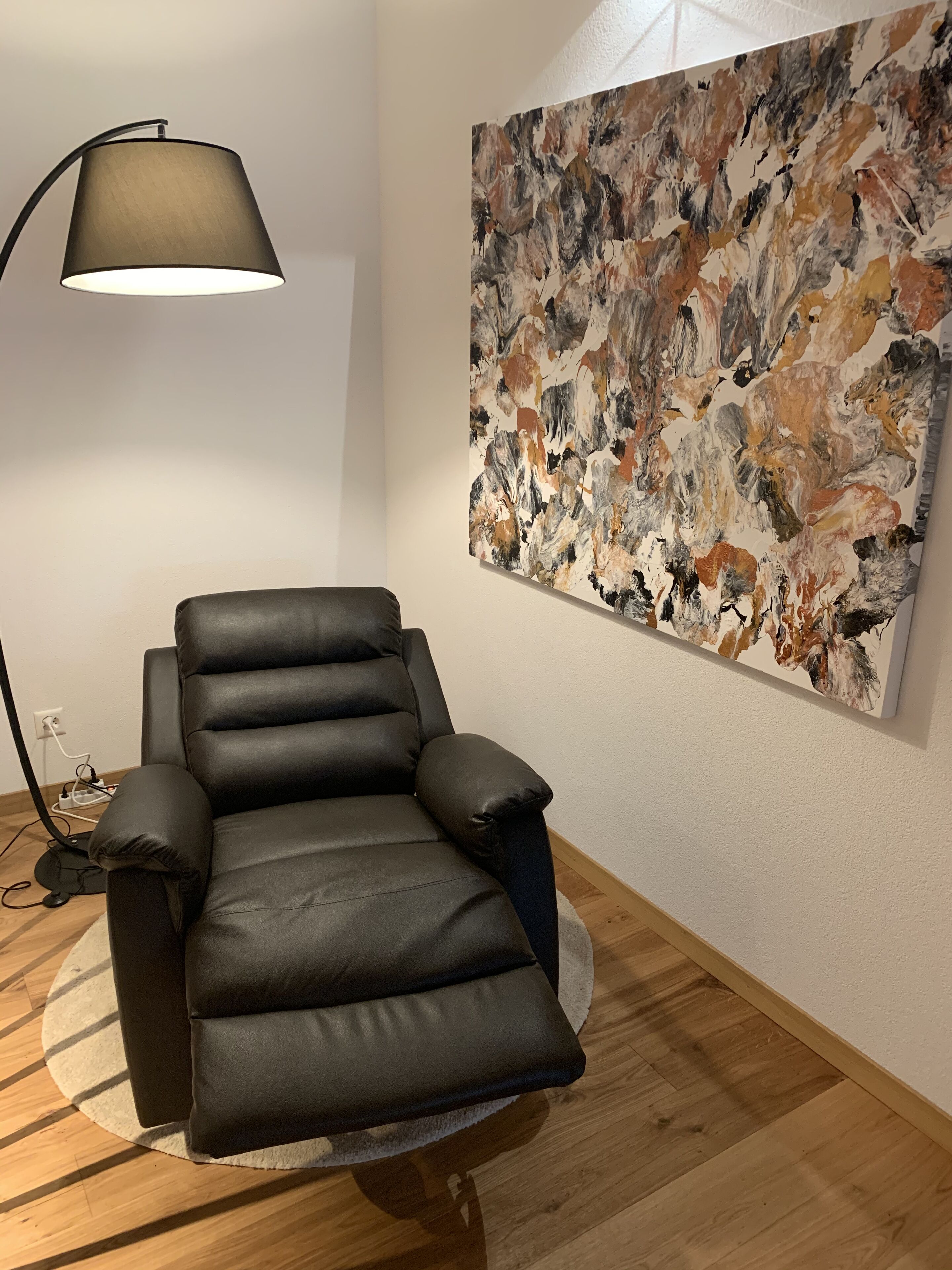 Una silla de cuero negro con reposapiés, una lámpara de pie y un cuadro abstracto grande en la pared.
