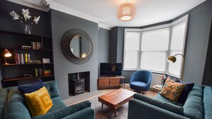 Ein modernes Wohnzimmer mit türkisfarbenen Sofas, einem Holztisch, einem Bücherregal, einem Kamin, einem Fernseher und einem großen Fenster mit Tageslicht.