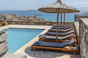 A poolside area with lounge chairs and a thatched umbrella, overlooking a rocky coastline and the sea.