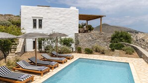 A pool area with lounge chairs and umbrellas, a white building with a stone wall, and a view of the landscape. - Indigo 4 Bedroom Villa Sea View Private Pool (Paros)