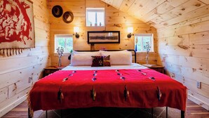 A cozy cabin bedroom with a sloped ceiling, wooden walls, a red bedspread, two bedside lamps, a framed picture, and a window with a view of greenery. - Farm - Animals - Barn - Gazebo - Fire Pit - 2xKing (Willington)