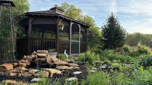 A wooden gazebo with a glass enclosure, surrounded by a garden with various plants and a small rock feature. - Farm - Animals - Barn - Gazebo - Fire Pit - 2xKing (Willington)