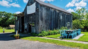 A rustic wooden barn with a large open door, a covered outdoor seating area with blue chairs, and a grassy lawn. - Farm - Animals - Barn - Gazebo - Fire Pit - 2xKing (Willington)