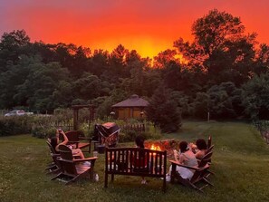A group of people seated around a fire pit in a garden with a gazebo and trees in the background. - Farm - Animals - Barn - Gazebo - Fire Pit - 2xKing (Willington)