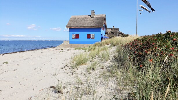 Ein Küstenhaus mit blauer Fassade und einem Reetdach, direkt an einem Sandstrand mit freiem Blick auf das Meer gelegen.