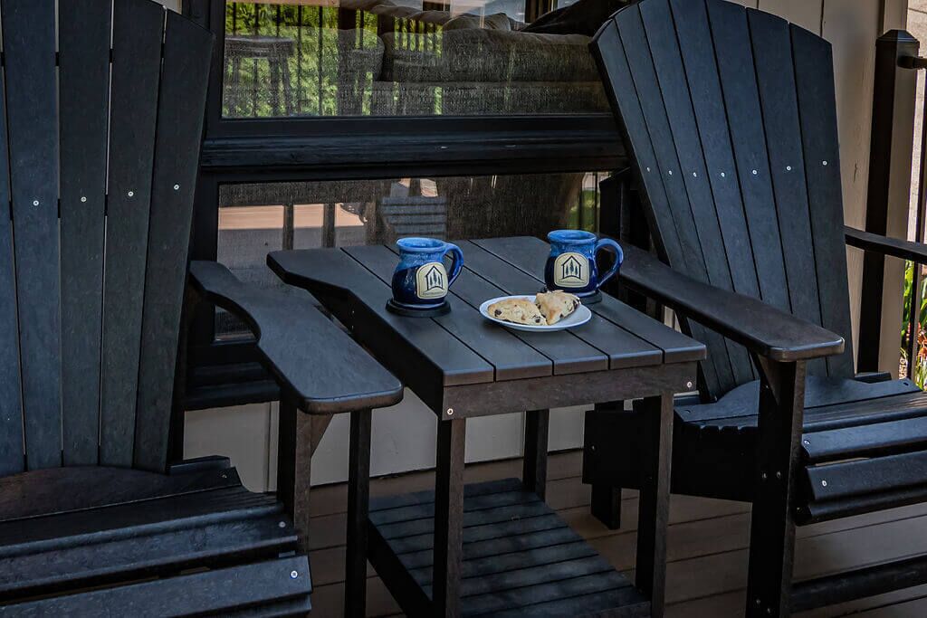A wooden table with two blue mugs and a plate of food, surrounded by black Adirondack chairs.