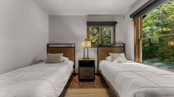 A bedroom with two beds, a nightstand with drawers, a lamp, and a window with a view of greenery.