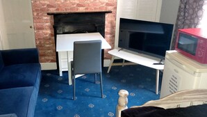 A living room with a blue sofa, a small table, a TV, a microwave, and a brick fireplace. - Phoenix Guest House (Barrow-in-Furness)