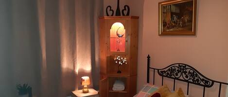 A bedroom with a bed, a nightstand, a wooden cabinet with a mirror, a painting on the wall, and a window with curtains.