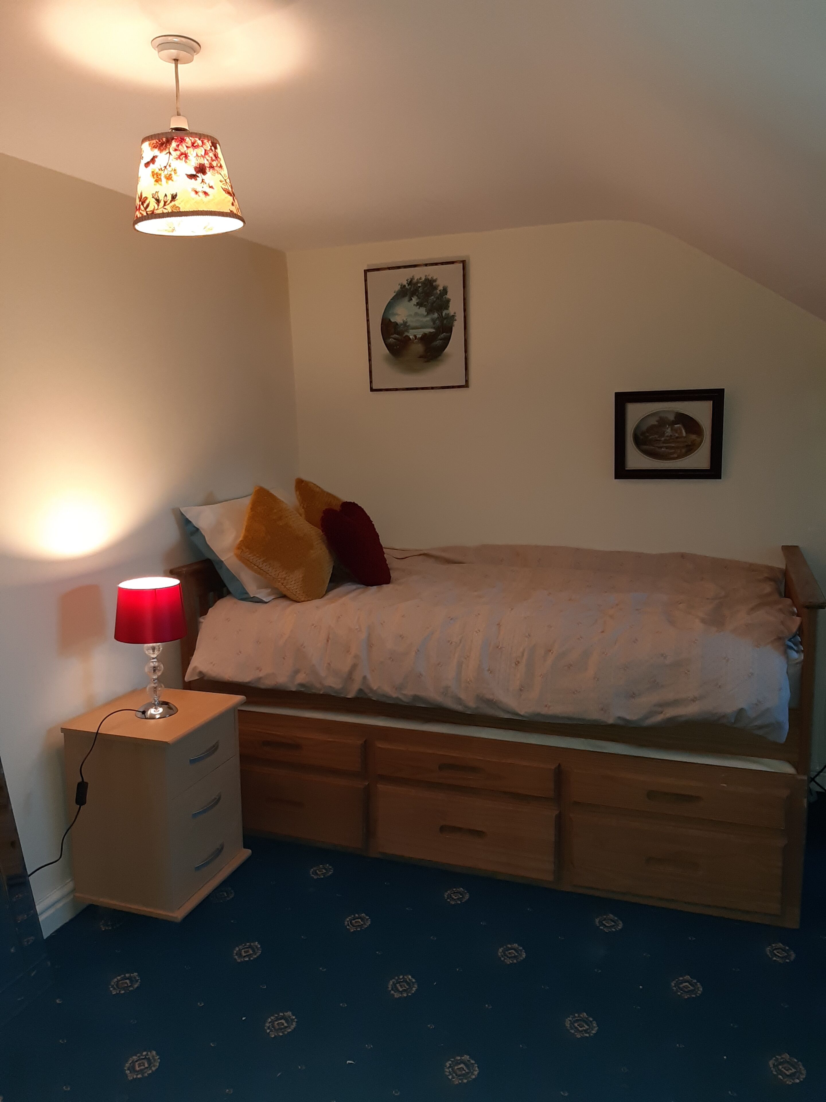 A bedroom with a bed, a bedside table with a lamp, a dresser, and two framed pictures on the wall.